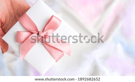 Female hand holding White gift box with pink ribbon on white silk texture background. Colorful bird feather. Holiday concept. Present for Valentines Birthday Mothers Womans day