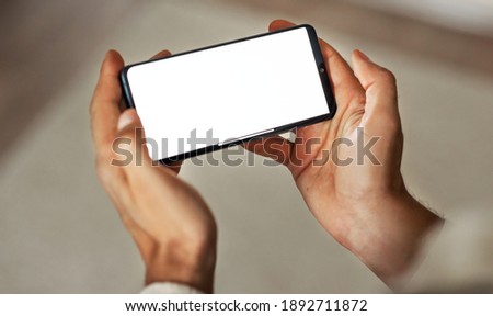 Closeup mockup image with blurred background of a man's hands holding modern mobile phone with blank white screen in a cozy room.