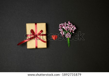 Flat lay. Valentine's Day. In the center is a gift box wrapped in craft paper and tied with a ribbon, a bouquet of lilac flowers. Between them is a small red heart. Black background.