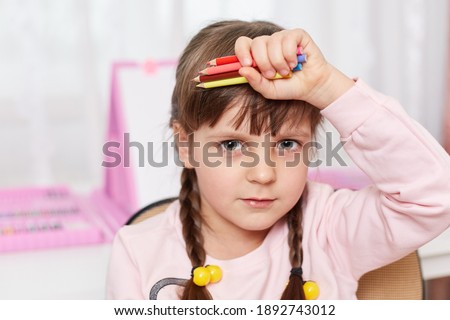 Cute little preschooler kid being tired of drawing, holding her pencils in hand, keeping her palm near forehead, looking sad, looks at camera, child painting long hours.