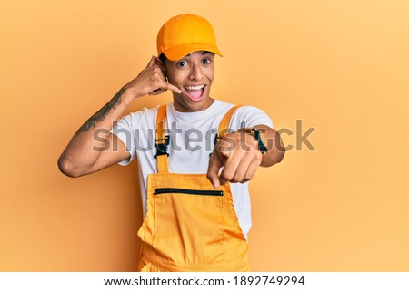 Young handsome african american man wearing handyman uniform over yellow background smiling doing talking on the telephone gesture and pointing to you. call me. 