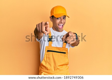 Young handsome african american man wearing handyman uniform over yellow background pointing to you and the camera with fingers, smiling positive and cheerful 