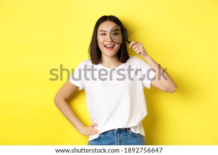 Cheerful asian girl searching for you, looking through magnifying glass and smiling, found something interesting, standing over yellow background