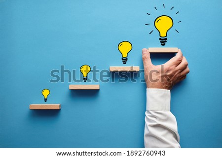 Hand drawn light bulbs representing a developing new idea on wooden steps with male hand placing the last step. Idea development, business vision or entrepreneurship concept. Royalty-Free Stock Photo #1892760943