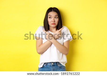 Indecisive asian girl need help with choice, pointing fingers sideways and looking confused, standing over yellow background