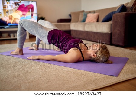Dedicated sportswoman lifting her hips and doing glute bridge exercise while following instructions of her fitness instructor on a TV at home.  