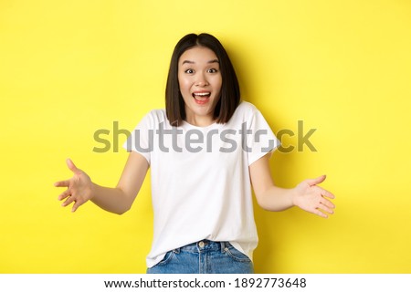 Excited young asian woman spread out hands as if holding large, big object, showing size and look amazed, standing over yellow background