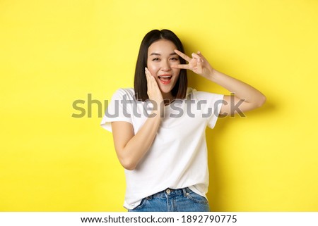 Lovely asian girl in white t-shirt posing with hand on cheek, showing peace sign on eye, standing over yellow background