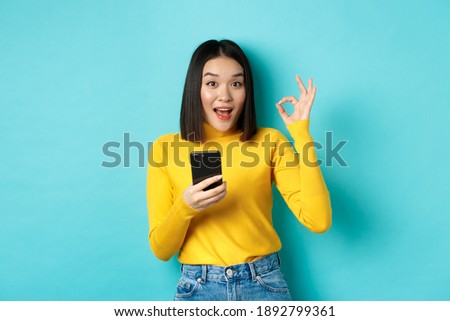 E-commerce and online shopping concept. Portrait of asian woman showing OK sign and using mobile phone, recommend application, standing over blue background