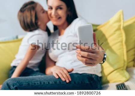 Daughter kissing smiling mother during selfie at home on blurred background