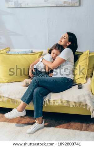 Full length of happy mother with closed eyes hugging daughter while sitting on couch