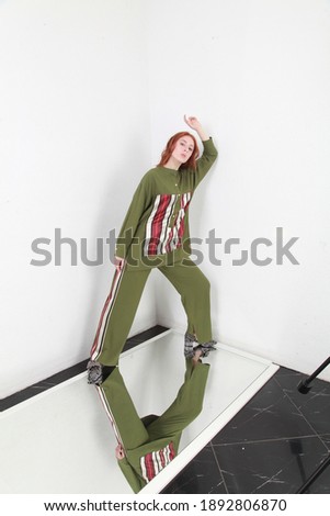 Full length of female ginger model posing in trendy altheisure style pants suit, posing standing over white walls background. Copy space for text. Catalog fashion photo shoot. Image in high key.
