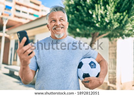 Middle age hispanic grey-haired man using smartphone and holding soccer ball at the city.