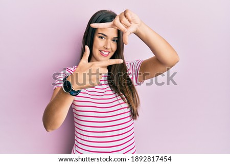 Young brunette woman wearing casual clothes over pink background smiling making frame with hands and fingers with happy face. creativity and photography concept. 