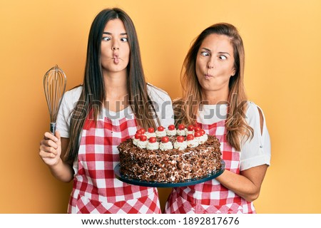Hispanic family of mother and daughter wearing baker apron holding homemade cake making fish face with mouth and squinting eyes, crazy and comical. 