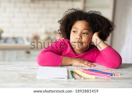 Boredom. Cute little bored black girl sitting at table with notebook and pencils in kitchen, thoughtful african american female child resting head on hand and looking away, feeling lonely, closeup
