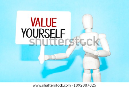 The wooden man holds a white sign with the text value yourself in his hands. The content of the lettering has implications for business concept and marketing.