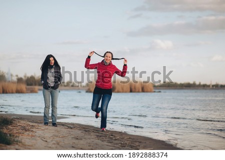 Two young girlfriends in blue jeans, a jacket and a shirt walk along the river bank in the evening at sunset. One of them is jumping on one leg, holding her braids with her hands and laughing happily.