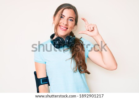 Beautiful young caucasian woman wearing gym clothes and using headphones smiling and confident gesturing with hand doing small size sign with fingers looking and the camera. measure concept. 
