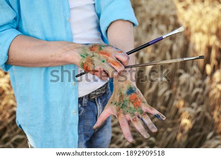 Close-up picture of dirty with paint hands, holding brushes, in front of yellow wheat field. Male artist wearing blue shirt and jeans painting in countryside. Outdoors artistic education activity.