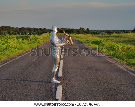 Man musician saxophonist in full body-hugging silver and silver electric suit holding golden alto saxophone, standing on empty road in summer with perspective. Freak, unusual person, alien, UFO.