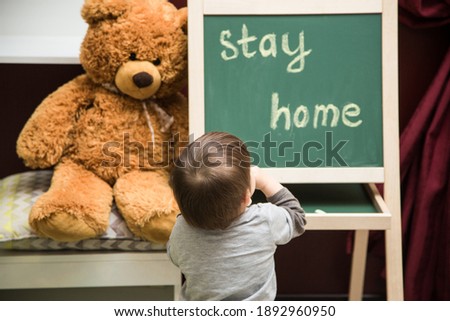 A teddy bear sits next to a children's blackboard on which a child toddler boy wrote "Stay home". Distance education symbol during the quarantine period due to the covid 19 coronavirus pandemic.