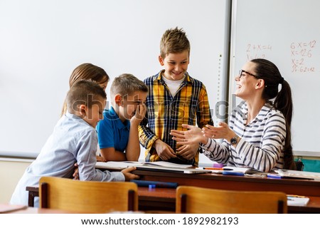 Female teacher helps school kids to finish they lesson.They sitting all together at one desk. Royalty-Free Stock Photo #1892982193