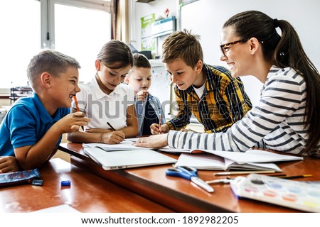 Female teacher helps school kids to finish they lesson.They sitting all together at one desk. Royalty-Free Stock Photo #1892982205