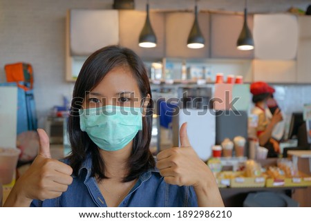 Young Asian woman or owner store wearing medical face mask during Covid19 outbreak in coffee shop to protect from Coronavirus, care for health and safety, showing good gesture sign. Royalty-Free Stock Photo #1892986210