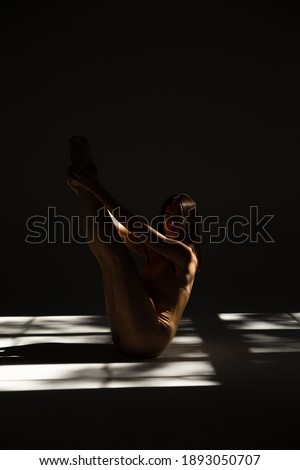 Silhouette of a girl sitting on a white background in a beige bodysuit. A slender girl in a beige bodysuit posing. Photo of a girl in bright light playing with shadows. Creative shot of a girl.