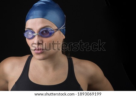 Young white girl with black swimsuit, blue cap and swim goggles, looks with a determined and concentrated air before the race, isolated on black background