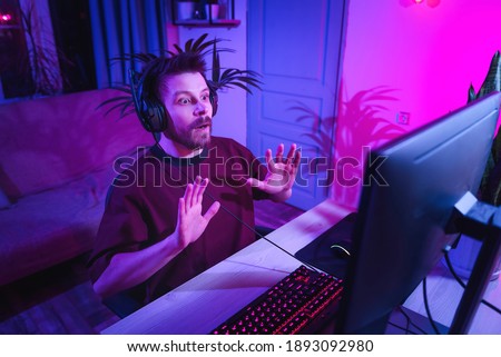Young gamer man playing video games scared in shock with a surprise face, afraid and excited with fear expression. Neon streamer studio. The guy with the headphones is having fun on the Internet.