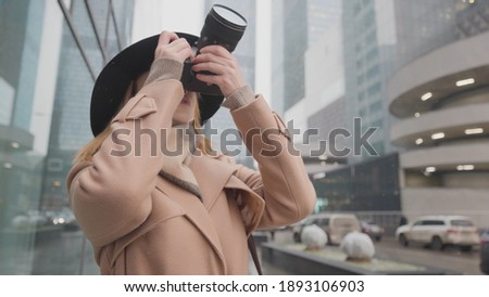 Beautiful woman takes urban winter landscape. Action. Stylish woman shoots urban landscapes on professional camera. Woman photographer takes modern urban high-rise buildings in snow