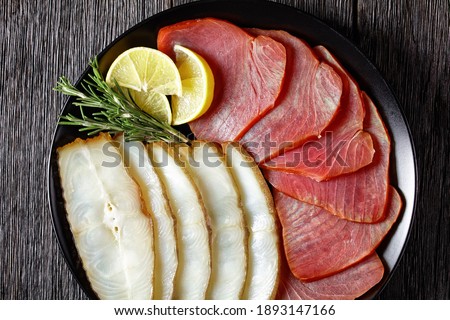 Smoked fish appetizer plate: cold smoked halibut and cold smoked tuna slices served on a black plate with rosemary sprigs and lemon wedges on a dark wooden table, top view, close-up