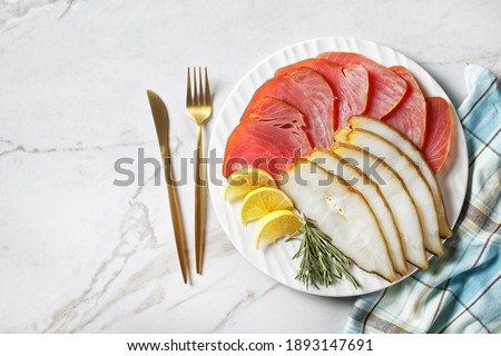 Smoked fish platter: cold smoked halibut and cold smoked tuna slices served on a white plate with rosemary sprigs, lemon wedges and English mustard on a light marble background, top view, close-up