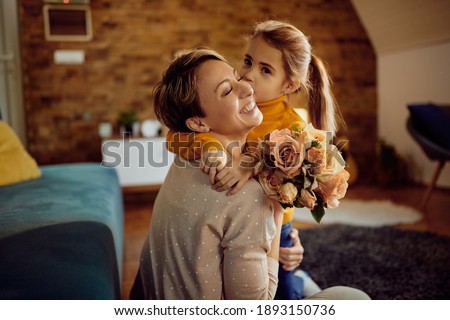 Happy woman receiving bouquet of flowers from her daughter who is kissing her at home.