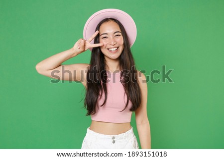 Smiling young asian woman 20s in casual pink clothes hat showing victory sign near eyes isolated on green wall background studio portrait. People sincere emotions lifestyle concept. Mock up copy space
