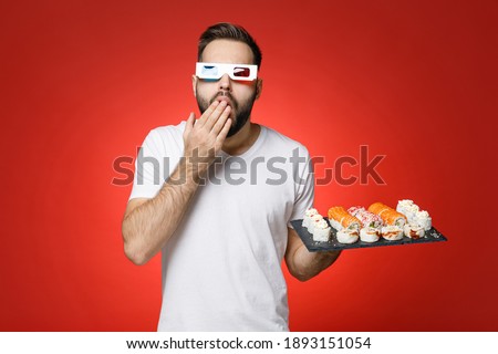 Shocked young man in white t-shirt 3d glasses cover mouth with hand watching movie film hold makizushi sushi roll served on plate traditional japanese food isolated on red background studio portrait