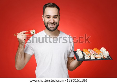 Smiling young bearded man 20s wearing casual white t-shirt hold sticks chopsticks makizushi sushi roll served on black plate traditional japanese food isolated on red color background studio portrait