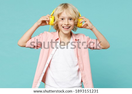 Excited little curly kid boy 10s years old wearing casual pink shirt listening music with headphones isolated on blue turquoise color background children studio portrait. Childhood lifestyle concept