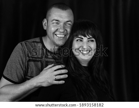 Black and white portrait of a couple in love in the studio,when a man hugs a woman shoulders from behind,both are standing,looking on the side and smilling.