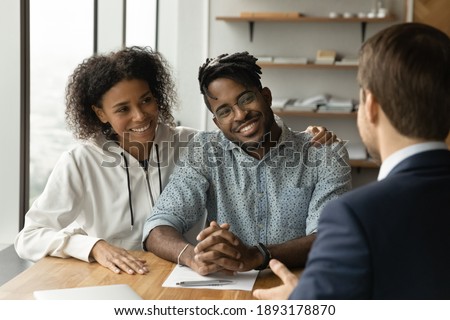 Smiling biracial man and woman talk with male real estate agent at office meeting. Happy African American young couple consult with realtor or broker, buy first shared house together. Rental concept. Royalty-Free Stock Photo #1893178870