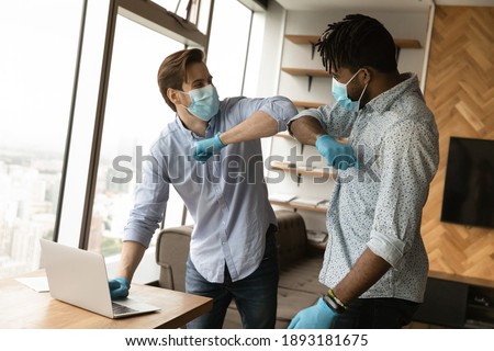 Smiling multiracial male colleagues in medical gear meet in office touch elbow greet during coronavirus pandemics. Diverse employees in face masks and gloves say hello at workplace. Covid-19 concept. Royalty-Free Stock Photo #1893181675