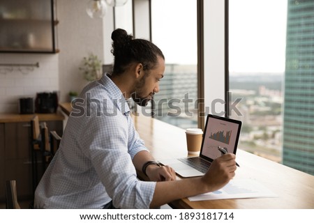 Focused African American male employee sit at desk in office work on computer analyzing graphs. Concentrated biracial man worker look at laptop screen busy with paperwork at workplace. Royalty-Free Stock Photo #1893181714