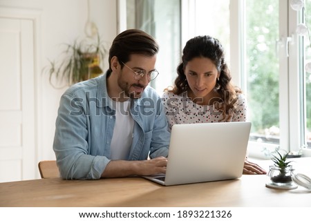 Young family using laptop together, sitting at wooden table in kitchen, attractive woman and smiling man wearing glasses surfing internet, shopping online, making purchases, booking tickets Royalty-Free Stock Photo #1893221326
