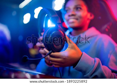 Headphones with microphone in hands of young african happy woman professional gamer online video game, neon background.