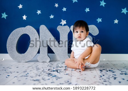 One year old baby boy in white t-shirt celebrates birthday near silver letters ONE on blue background with stars and silver sparkles