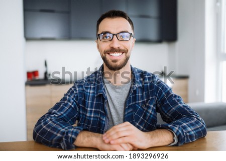 Webcam portrait of happy freelancer guy during video conference, he looks at the camera and smiling. Young adult stylish intelligent man wearing eyeglasses distantly works from home