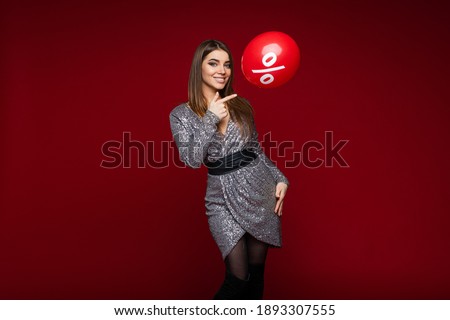 Smiling young girl in festive dress pointing on balloon with per cent sign on red background, sale and discount concept