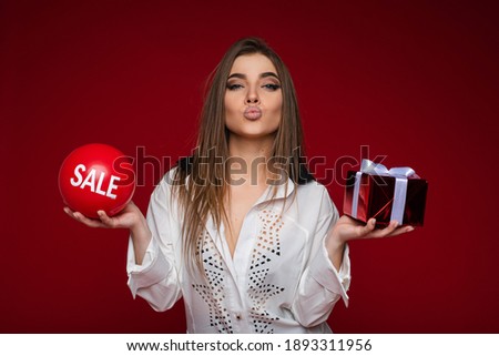 Beautiful young girl holding festive gift and balloon with sale lettering on red background with copy space for ad